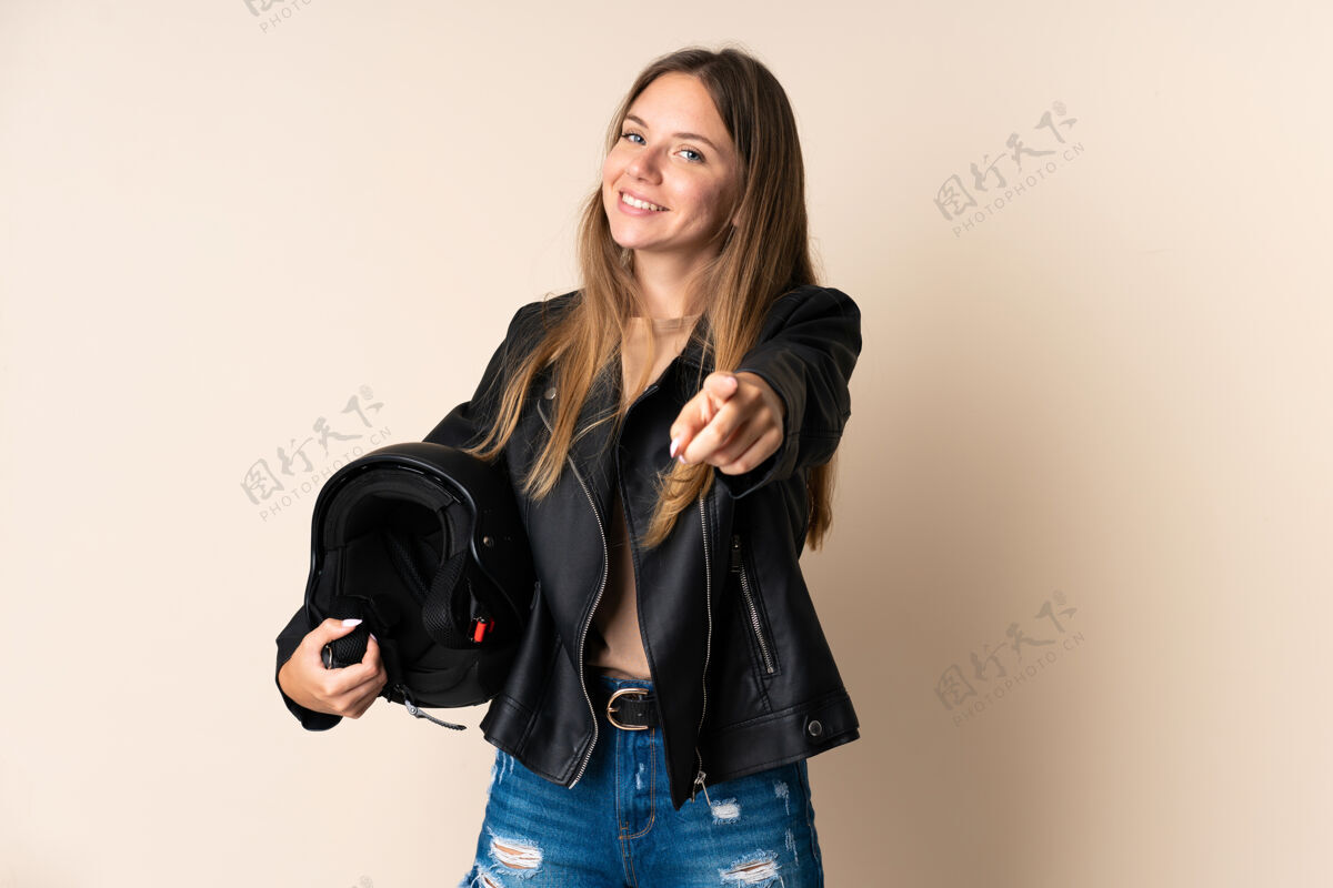 表情立陶宛年轻女子手持摩托车头盔 孤立地站在米色背景上 幸福地指着前方微笑女性司机