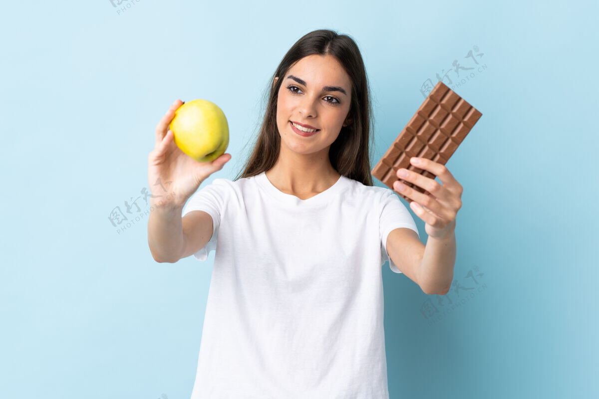 糖果一个年轻的白人妇女 一只手拿着巧克力片 另一只手拿着苹果 被隔离在蓝色背景下女孩年轻女人