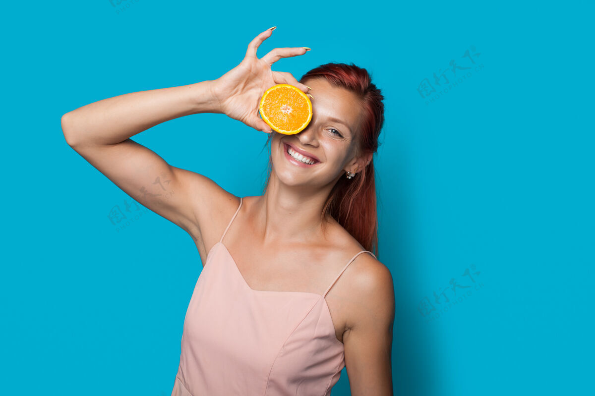 护理红头发的女人在蓝色的工作室墙上摆着一副柠檬片遮住眼睛的姿势皮肤柠檬女人