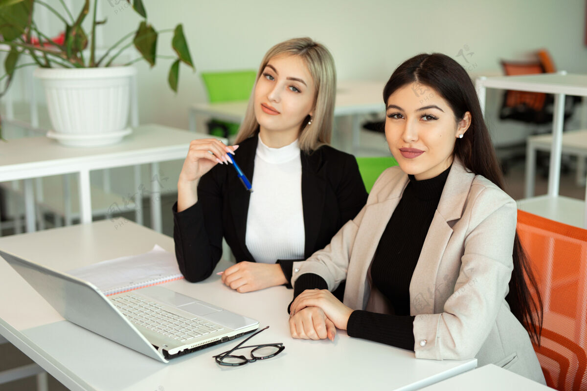 项目两个漂亮的年轻女子在办公室的桌边拿着笔记本电脑女性教育咨询