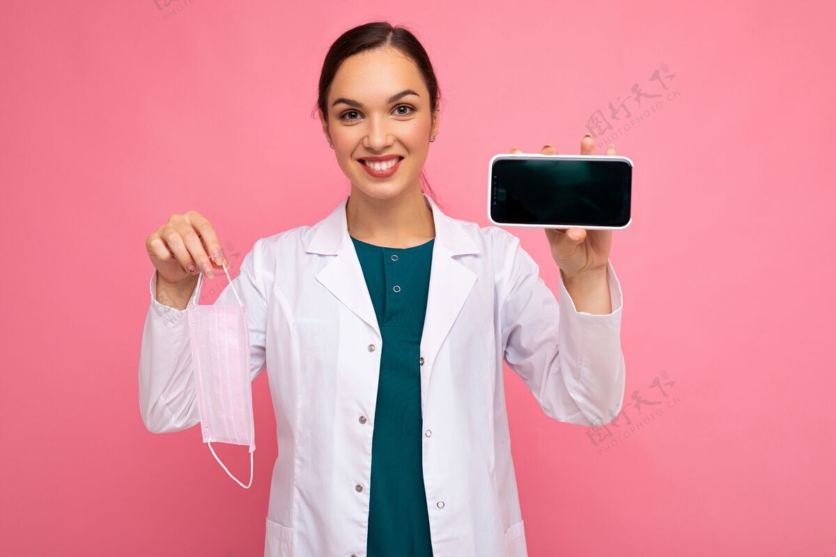 聪明迷人的微笑医学年轻女子身穿白大褂手持面具展示现代手机隔离在粉红色背景下看着摄像头.mock起来 切断通讯表情距离