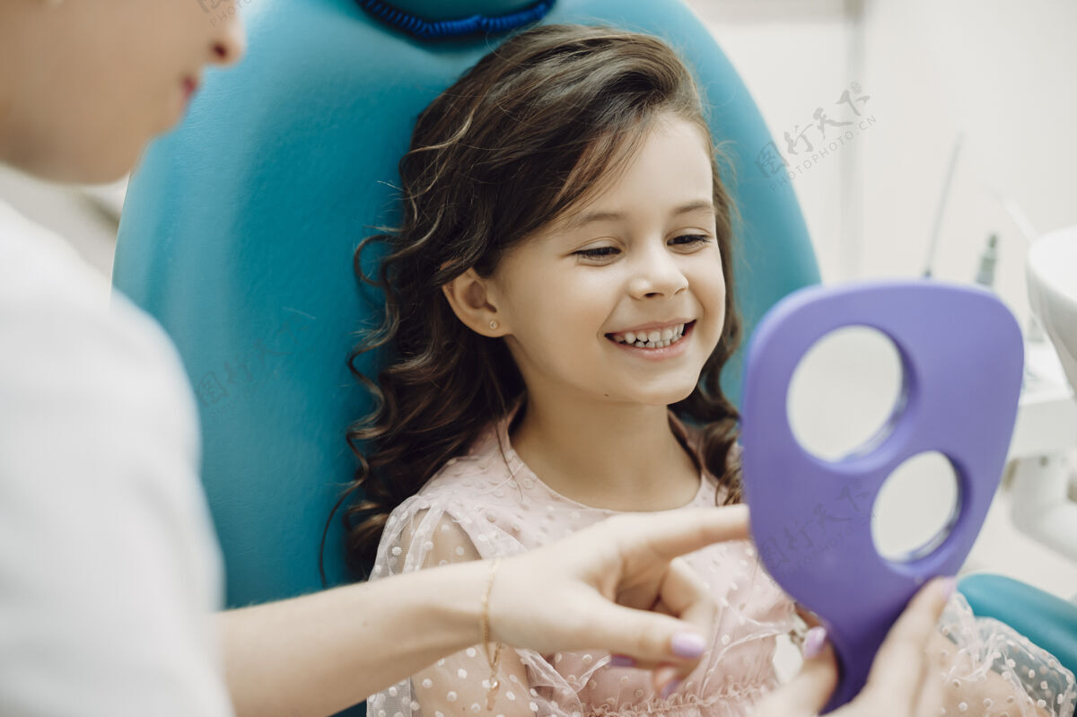 办公室一个可爱的小女孩坐在口腔科的座位上 在儿科口腔科做牙齿手术后看着自己的牙齿的画像初级帮助人