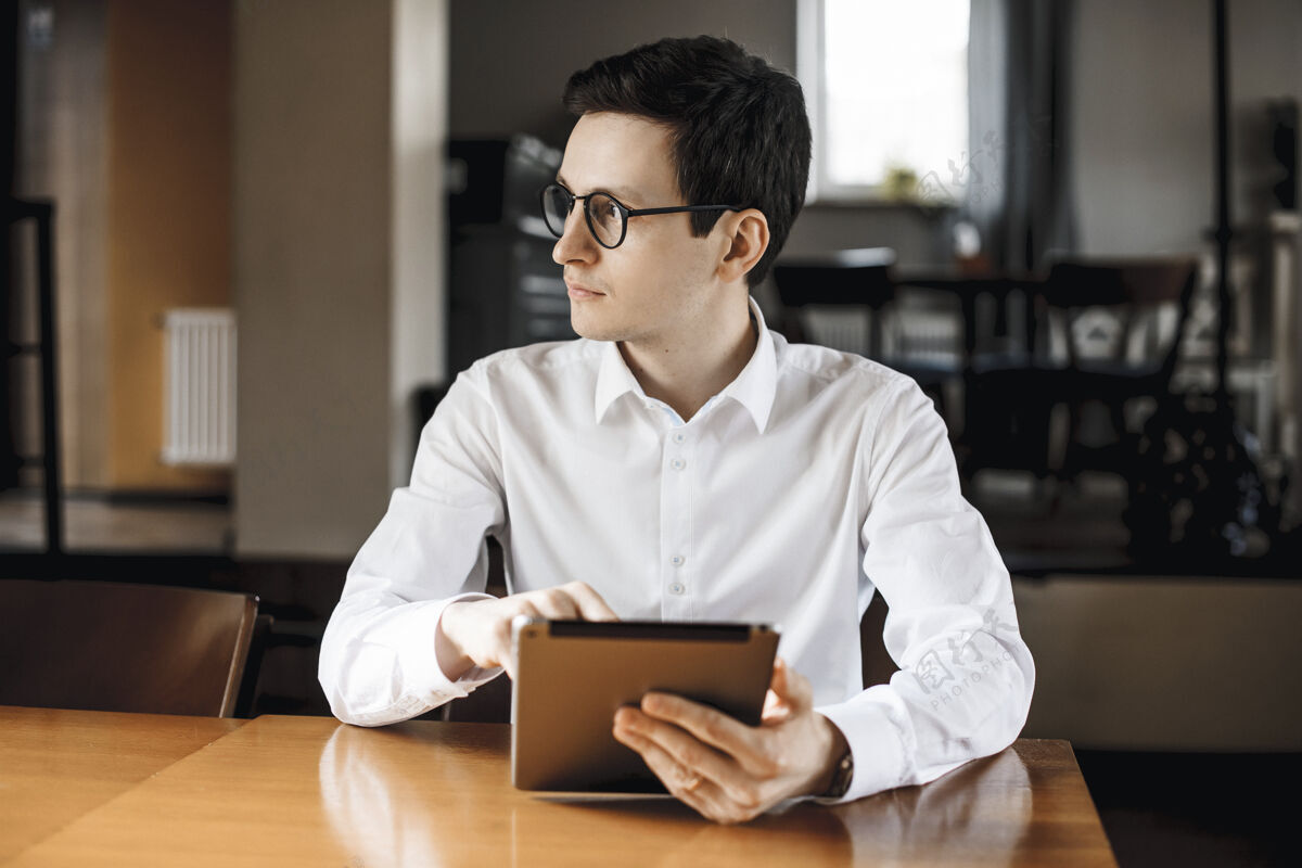 房间一个穿着白衬衫戴着眼镜的帅哥经理的画像 他拿着一个平板电脑坐在咖啡店的办公桌旁沉思办公室商务人士