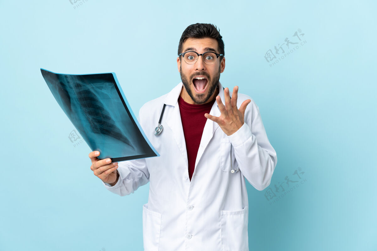 惊人专业的创伤科医生拿着蓝色背景上隔离的X光片 脸上露出惊讶的表情护理手势哇