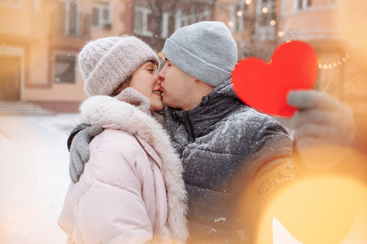 霜冻情人节的概念 相爱的情侣在冬天的雪地里亲吻拥抱帕克·杨男人拿着一个红色的纸心和他的女朋友一起庆祝情人节一对情侣在一起感觉很温暖约会给予惊喜