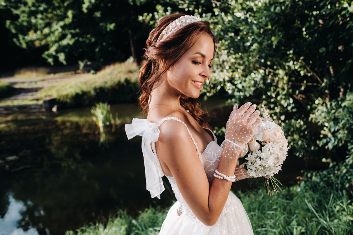 水一个优雅的新娘在一个自然公园里穿着白色礼服 手拿花束的肖像爱金发花