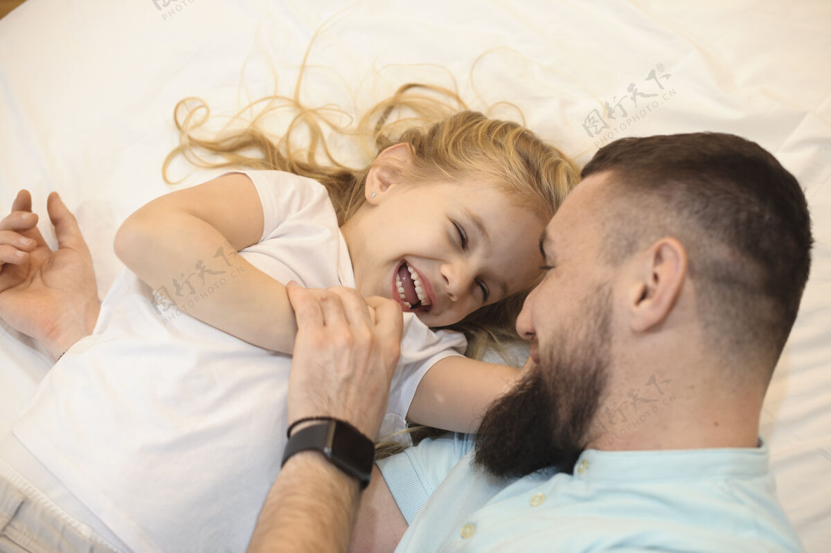 可爱一个可爱的金发小女孩在床上和她年轻的留胡子的父亲玩耍时闭着眼睛笑的画像金发教育空闲时间