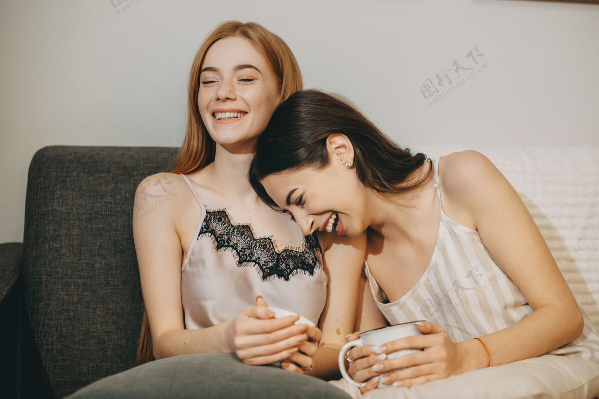 牙关一个可爱的红发女人笑的肖像 而她的女性朋友正靠在她的肩膀上笑着闭着眼睛坐在沙发上沙发沙发室内