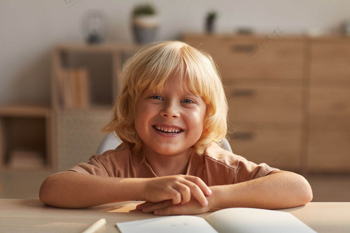 看快乐的金发小男孩坐在桌旁拿着文案微笑的画像天真坐着金发
