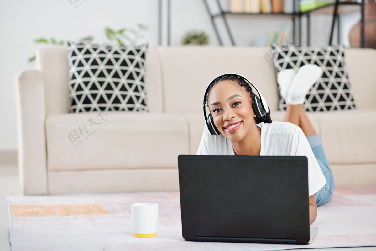 编程正面的年轻黑人妇女戴着耳机躺在地板上 在家里用笔记本电脑工作的肖像家庭电脑微笑