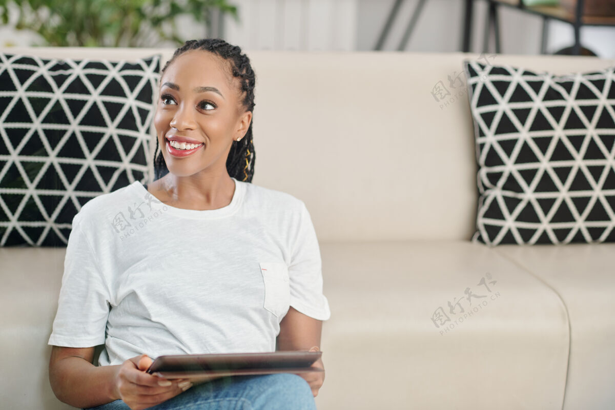 舒适可爱的年轻美籍非洲裔女子 戴着可怕的长发 坐在房间的地板上 手里拿着数字平板电脑 望着别处积极的情绪牙齿般的微笑女士