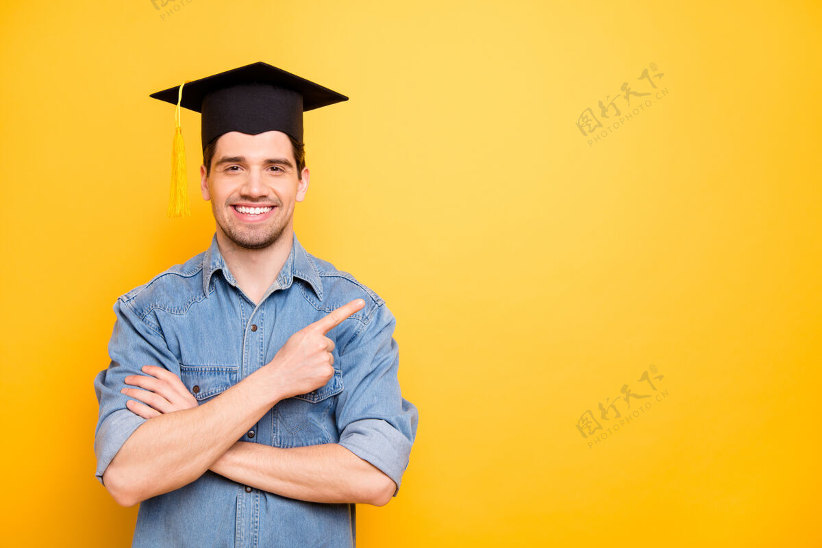毕业照片中开朗的正派帅哥微笑着牙齿指着空旷的空间双手合十戴着帽子隔离生动的彩色墙壁手指学士教育