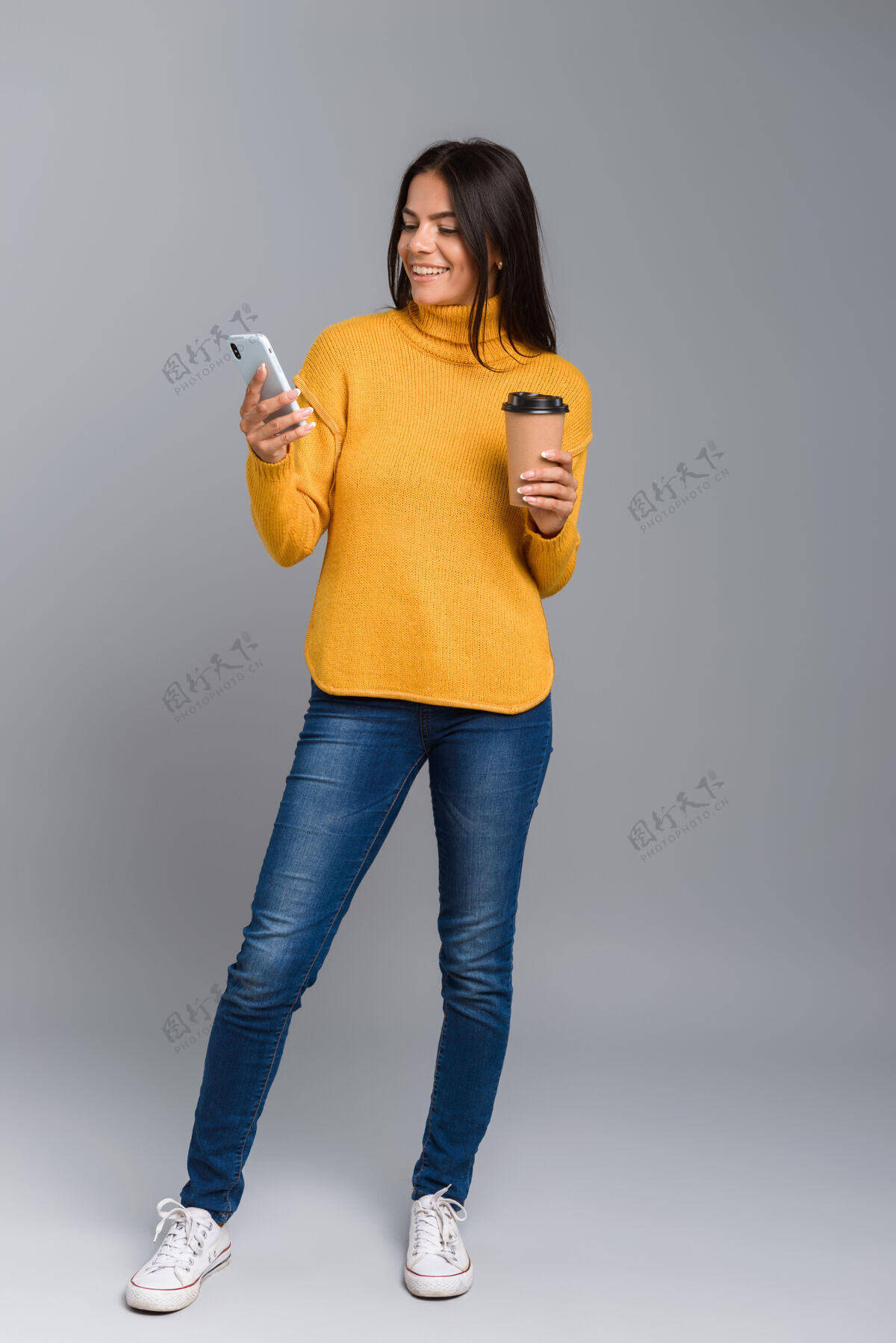白种人一个随意的年轻女子的全长肖像 隔着灰色的墙 拿着外卖咖啡用手机应用程序微笑女性