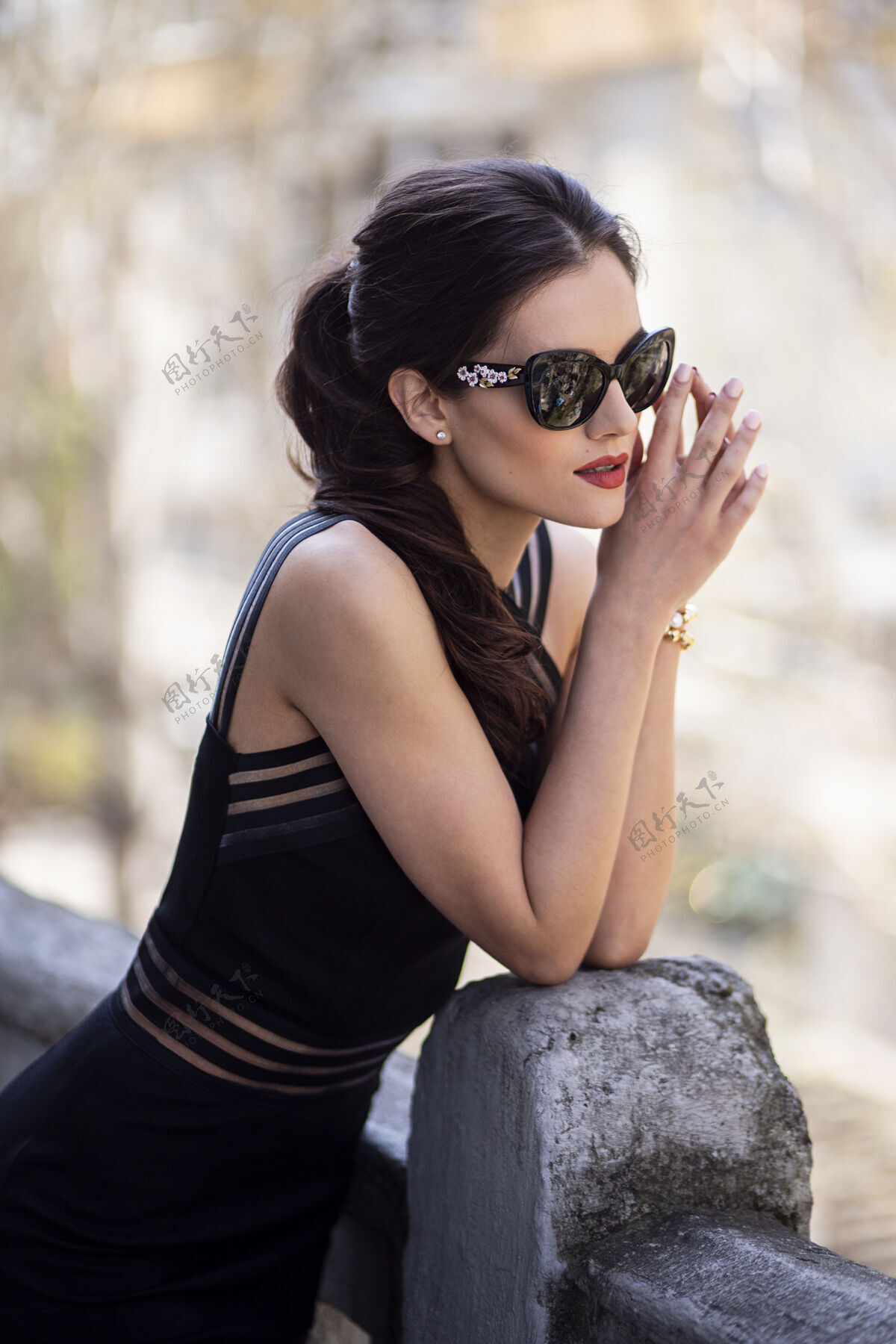 时尚一个美丽的 黑发的年轻女子戴着黑色太阳镜和裙子 梳着马尾辫 化着妆 在阳光明媚的日子里在阳台上摆姿势发型户外太阳镜
