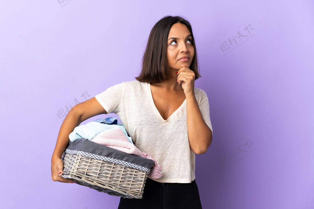 困惑年轻的拉丁女子拿着一个衣篮孤立在紫色的怀疑洗衣机失望湿
