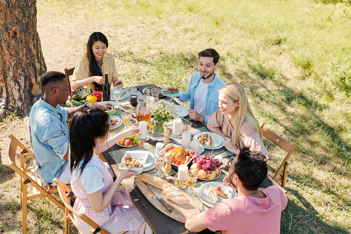 聚在一起一群年轻快乐的跨文化朋友聚在餐桌旁 在阳光明媚的日子 在松树下享用自制的户外晚餐父母欢呼纽带