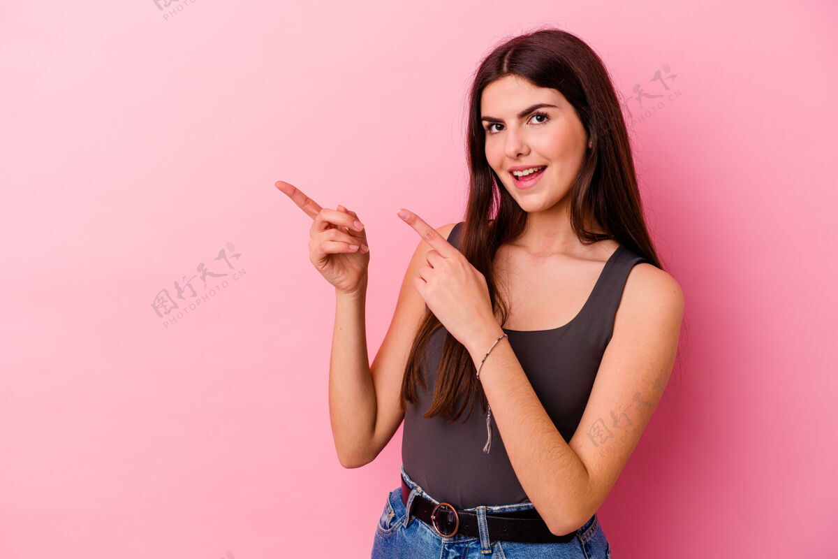 微笑年轻的白人妇女孤立在粉红色背景下兴奋地用食指指着女人年轻惊喜