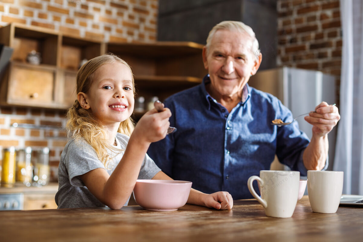 营养快乐可爱的小女孩坐在她的祖父 而享受早餐在厨房里成人退休房子