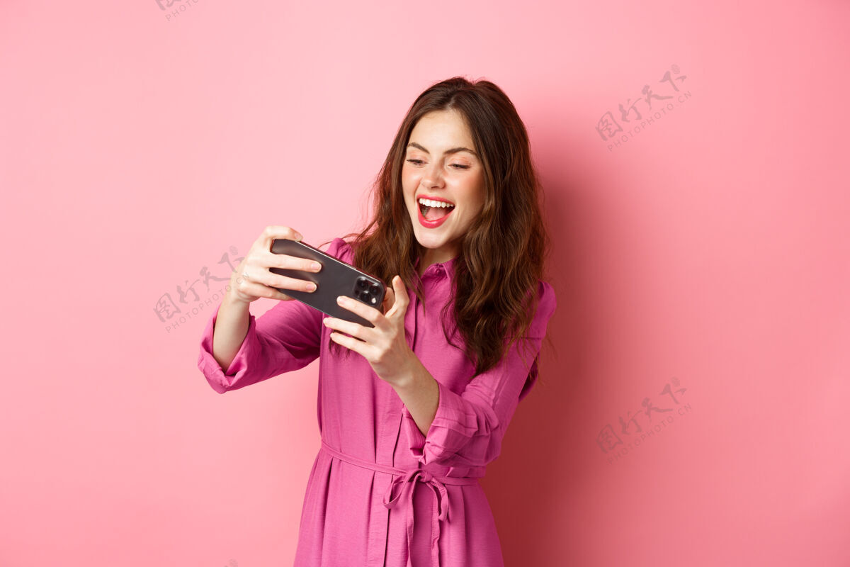 请科技概念无忧无虑的年轻女子倾斜身体 水平地拿着智能手机 在手机上玩电子游戏 站在粉红色的墙上复制空间咧嘴笑科技在线
