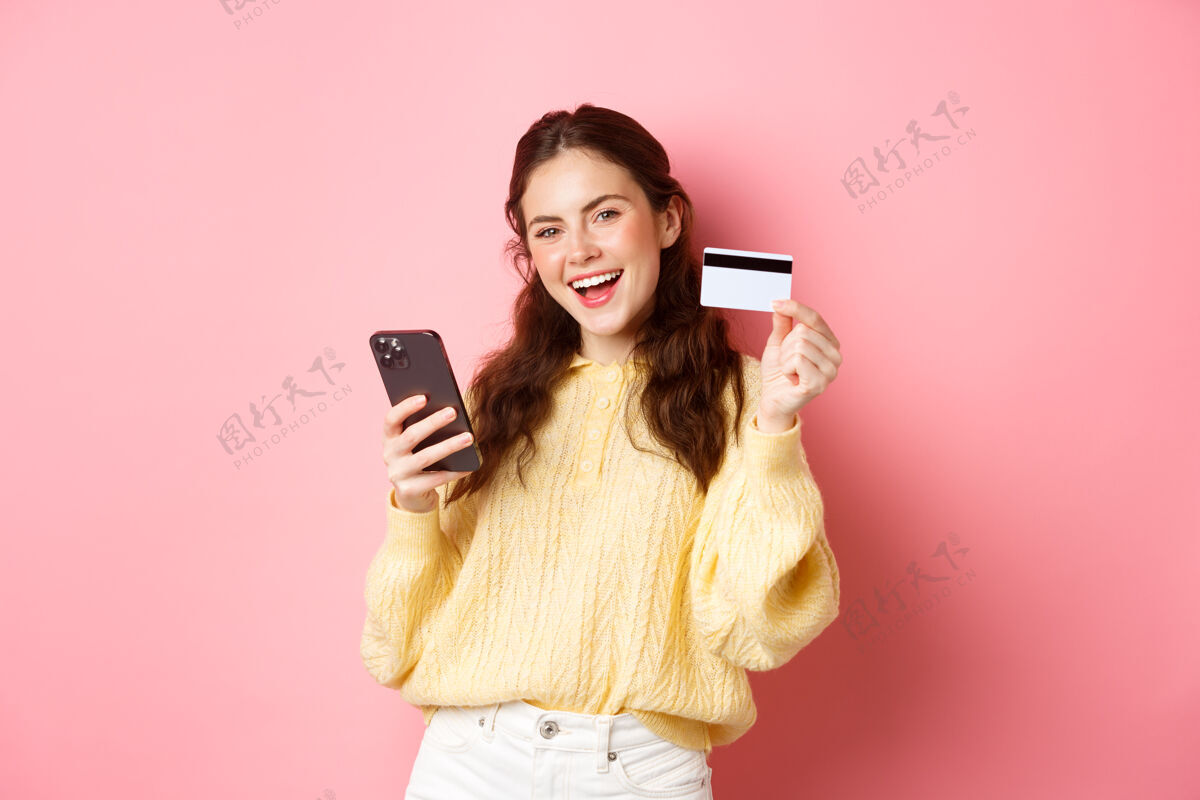 积极科技和网上购物快乐和满意的女性客户 展示塑料信用卡和使用手机应用程序在线支付 站在粉红色的墙应用程序魅力时尚