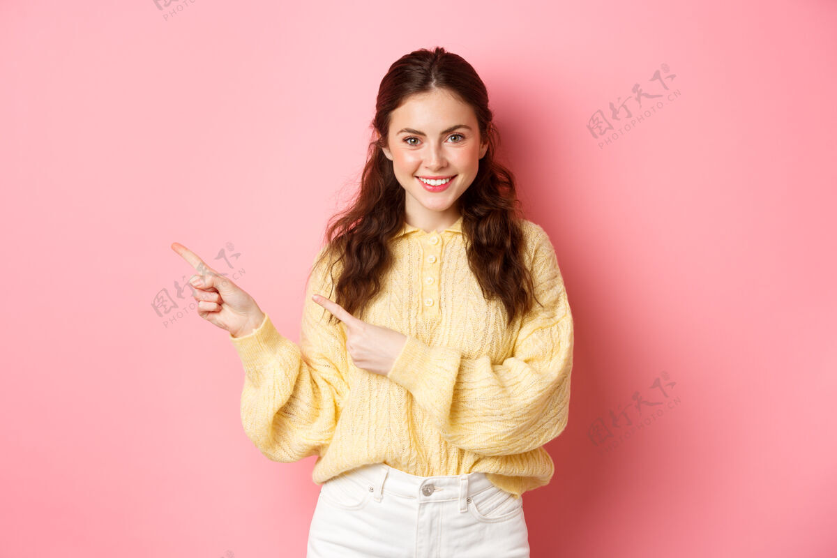 时尚微笑自信的女人用手指指着商标的肖像 展示了一张坚定的脸 站在粉红色的墙上的宣传文字学生女性微笑