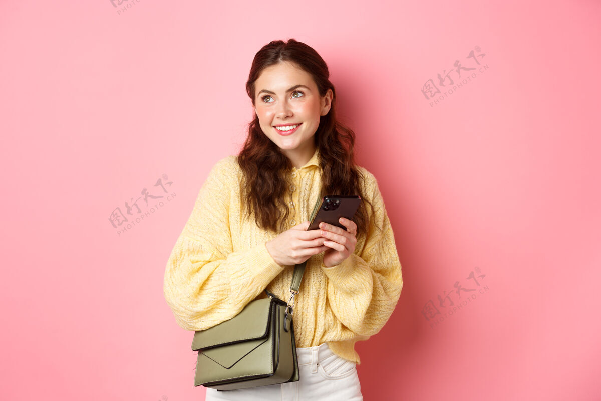 成人时髦的现代女孩 面带微笑 一边看一边浪漫 一边在智能手机上的约会软件上聊天 拿着钱包站在粉色的墙上时尚手势女性