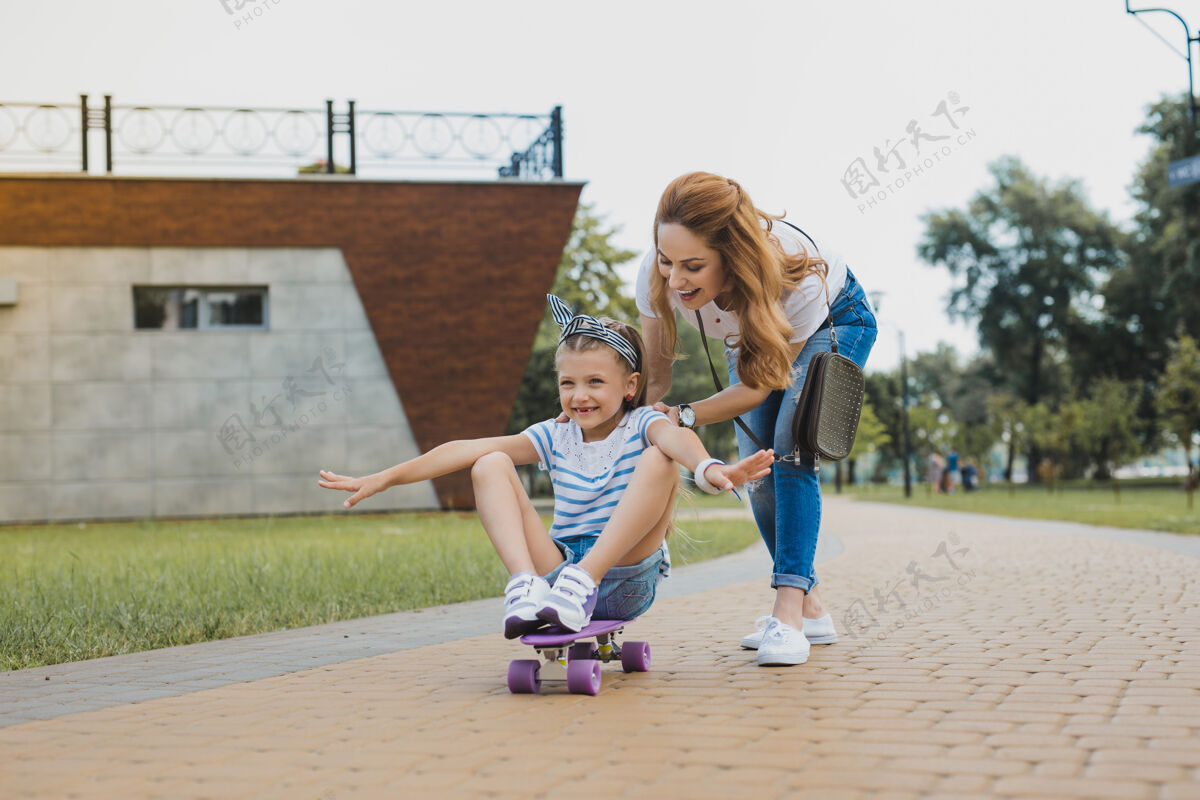 头发坐在滑板上兴奋有趣的女孩穿着时髦的发带坐在滑板上靠近她的母亲环境周末卡路里