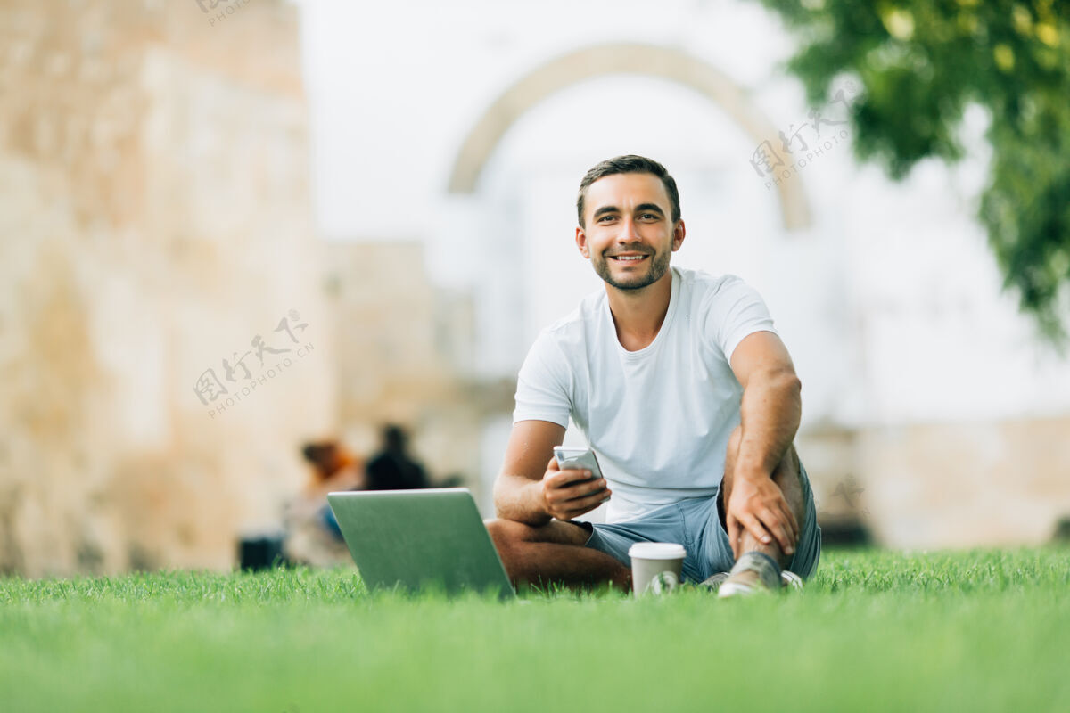 眼镜年轻人用他的笔记本电脑和咖啡去草地上工作清晰公园冲浪