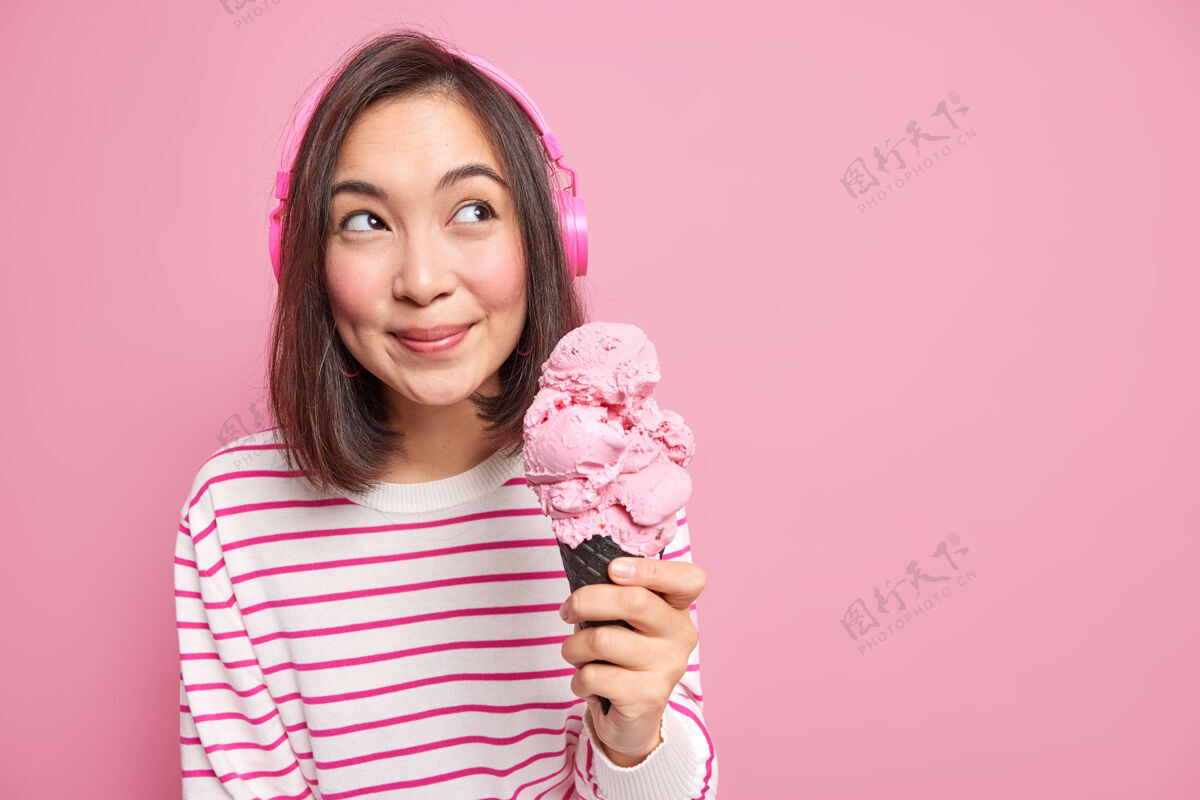 女性长着一头黑发的可爱的亚洲年轻女子拿着大大的草莓冰淇淋 若有所思地移开视线 戴着立体声耳机 隔着粉色的墙壁复制空间衣服模特立体声