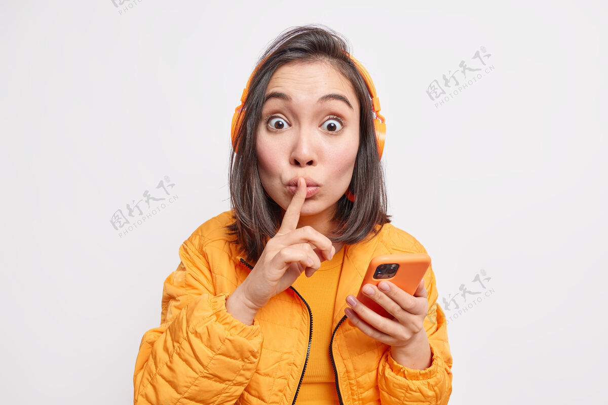 沉默拍到一个惊讶的女人在闲聊什么让沉默的手势告诉秘密举行现代手机使用耳机听音乐穿着时尚的橙色夹克不要告诉任何人八卦情感姿势
