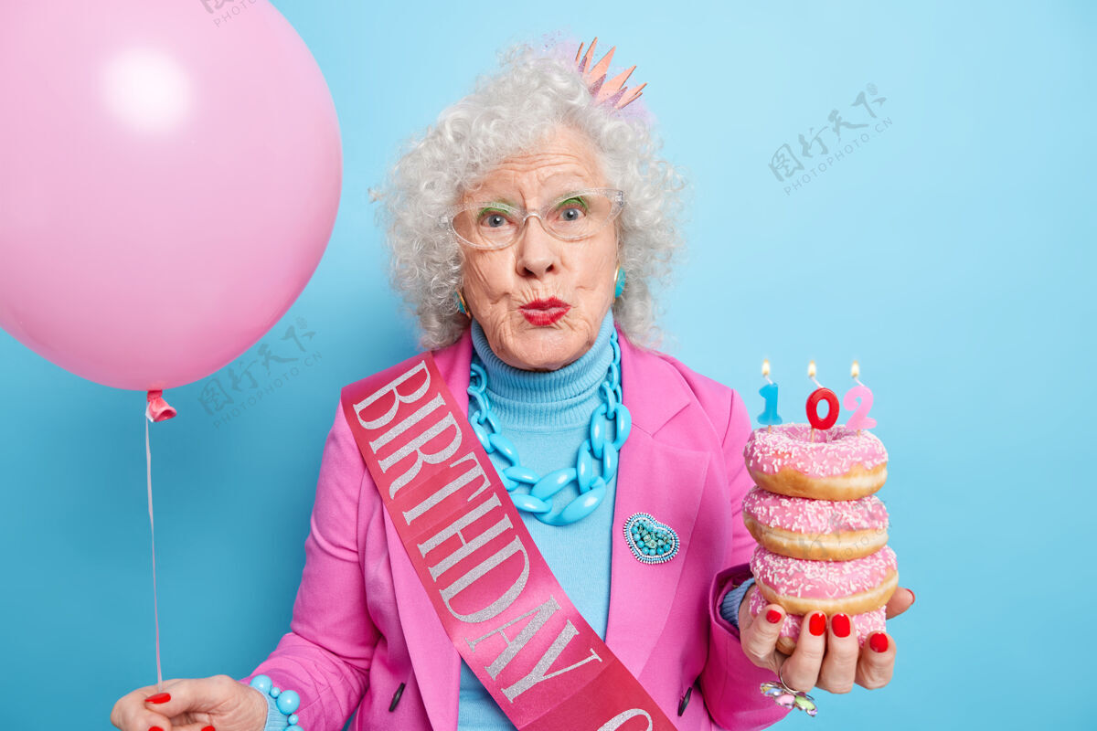 女人美丽的卷发老太太双唇紧闭享受生日庆典手持一堆美味的甜甜圈和蜡烛充气气球女士事件姿势