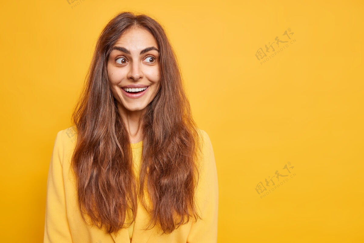 微笑快乐积极的女人留着长长的黑发微笑愉快地专注在一旁有着好奇快乐的表情模型对着鲜艳的黄色墙壁空白的复制空间为您提供信息情感概念工作空白反应