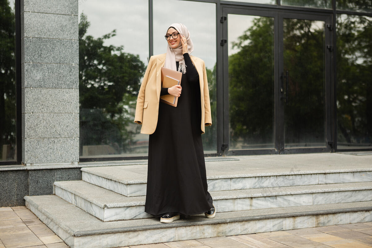 自营职业现代时尚的穆斯林妇女戴着头巾 穿着商务风格的夹克和黑色的长袍 带着笔记本电脑走在城市街道上民族职业女性