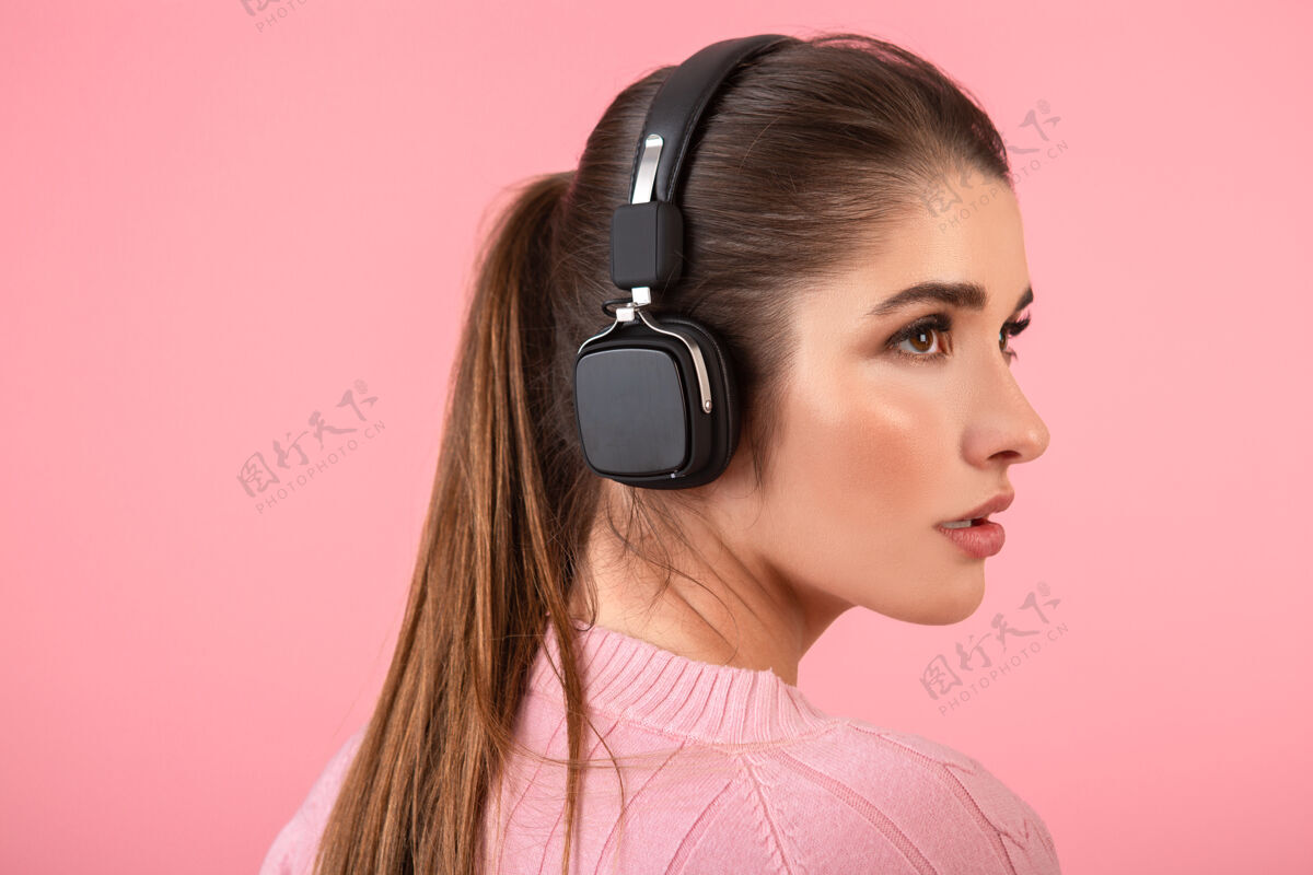 耳机年轻迷人的女人戴着无线耳机听音乐穿着粉色毛衣微笑着快乐积极的心情摆在粉色背景上欢乐漂亮音频
