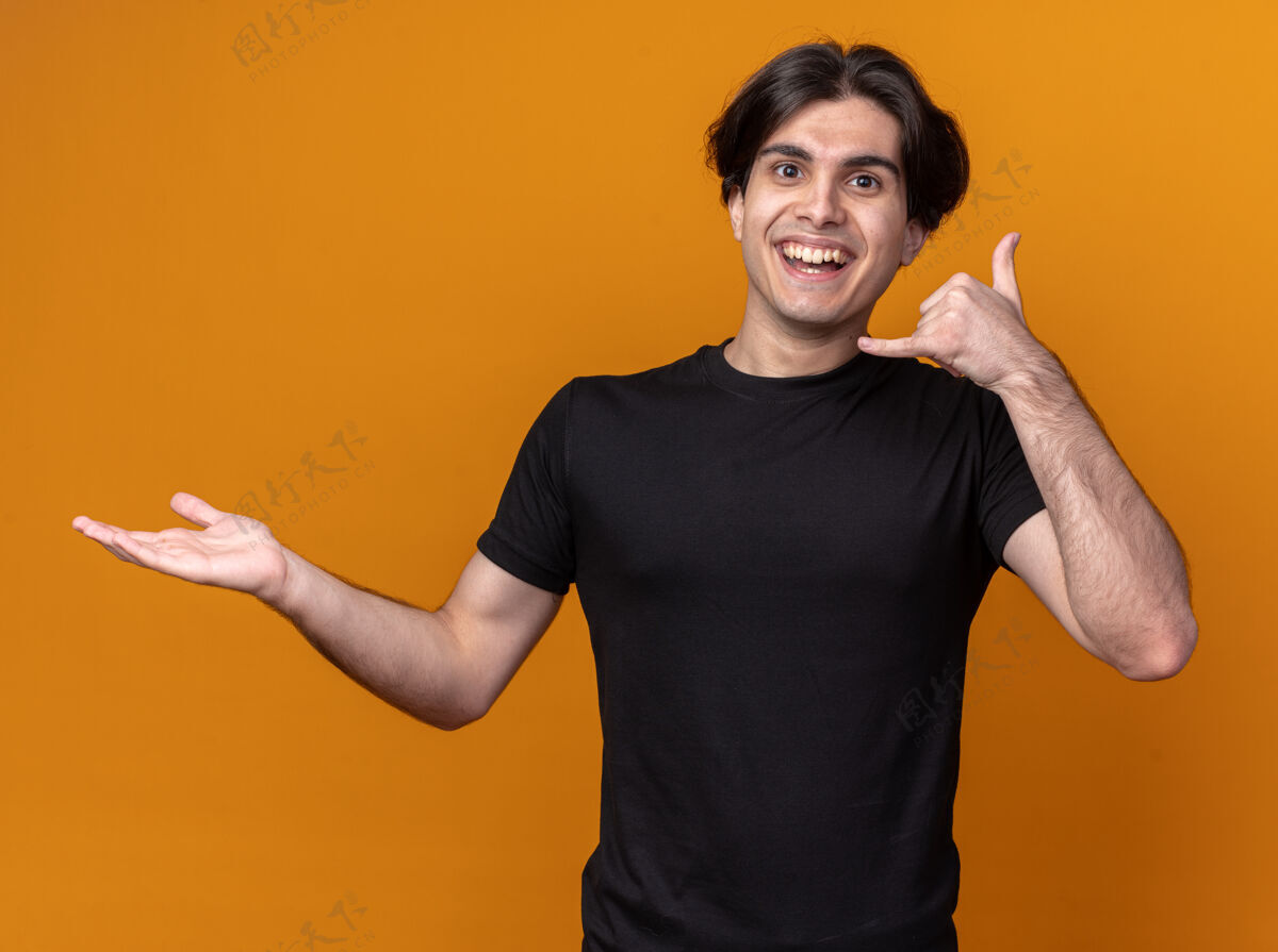 年轻带着微笑的年轻帅哥穿着黑色t恤展示着打电话的手势摊开手孤立在橙色的墙上 留着复印空间站着脸人