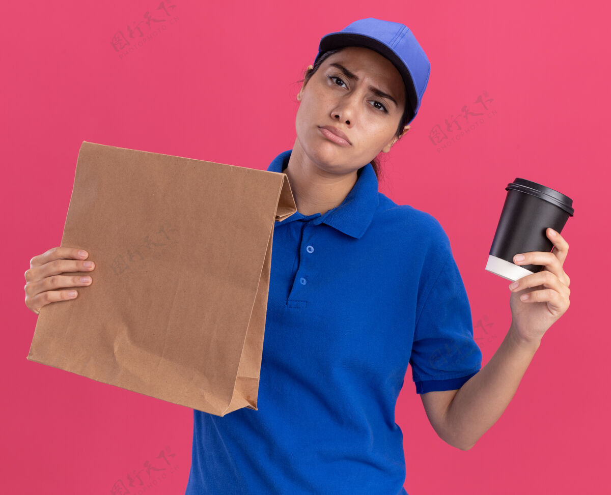 制服年轻的送货员穿着制服 戴着帽子 手里拿着一个纸食品包 粉红色的墙上隔着一杯咖啡帽子食物递送