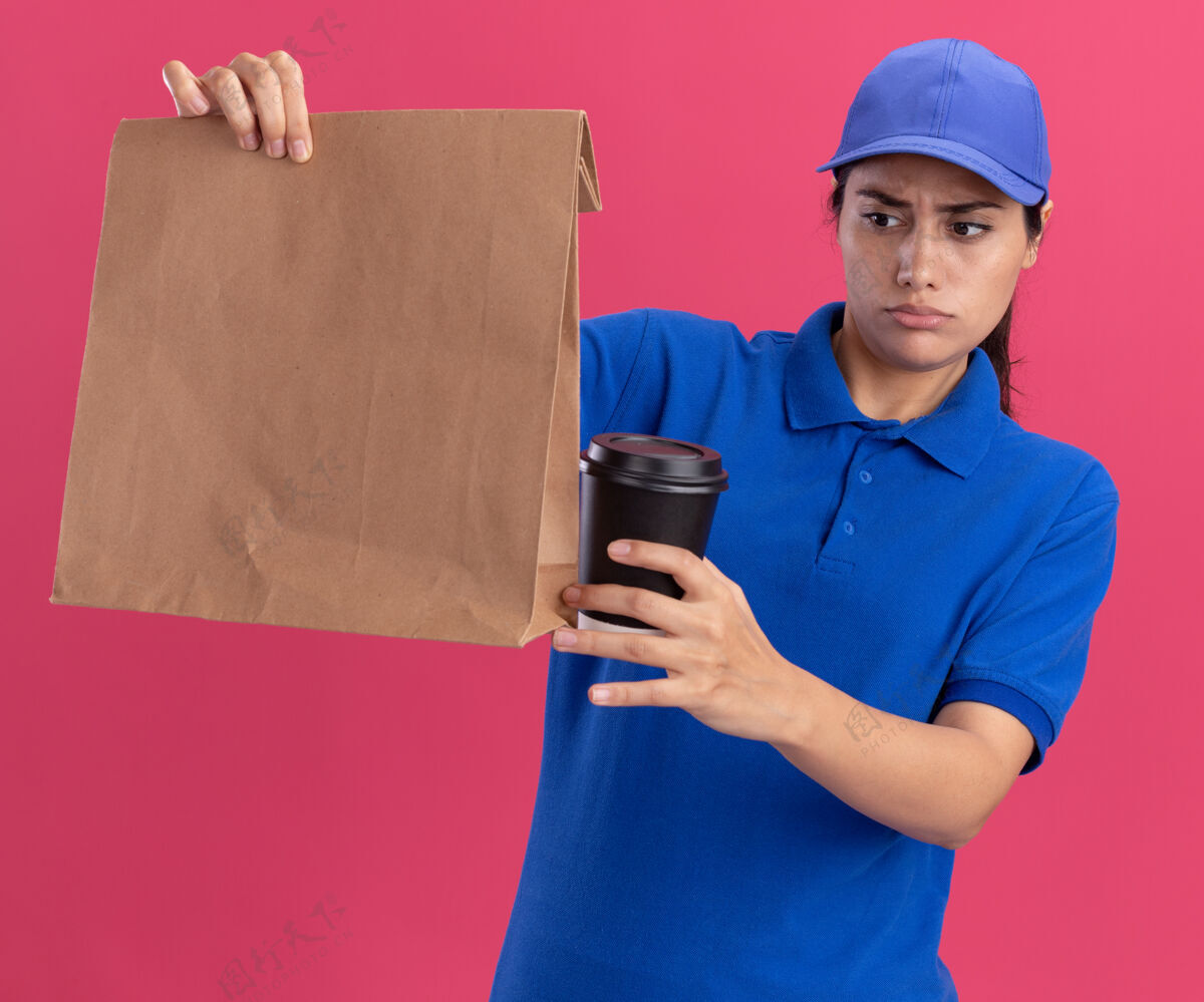 穿着困惑的年轻送货女孩穿着制服 戴着帽子 看着纸食品包装 粉红色的墙上隔着一杯咖啡杯子表情咖啡