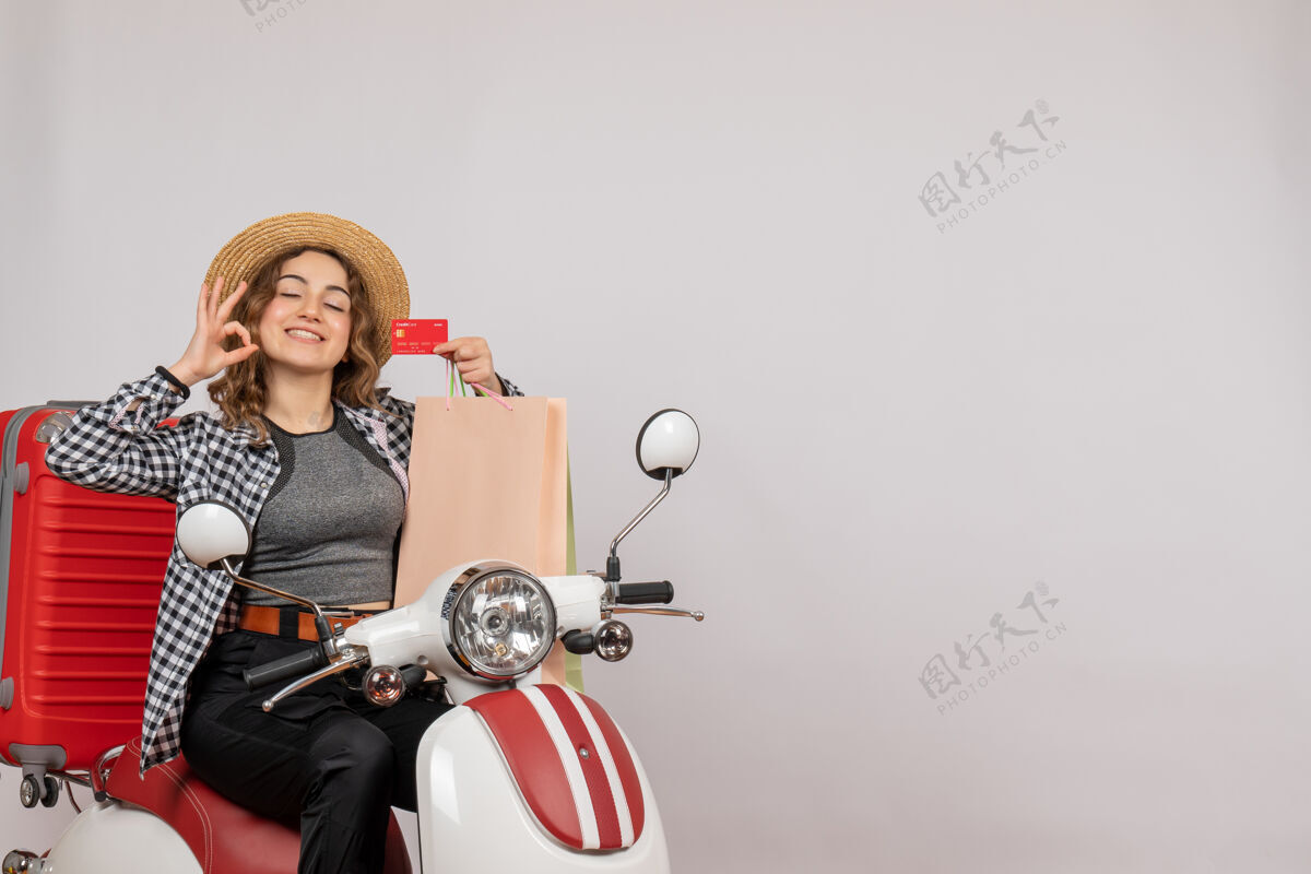信用卡正面图：骑着轻便摩托车的年轻女子拿着卡片做着“ok”标志坐着灰色成人