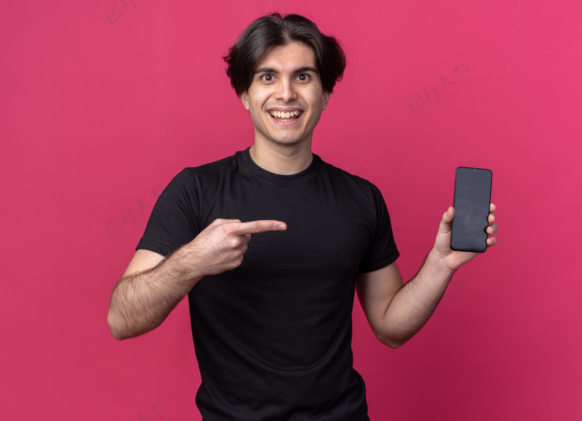 男人面带微笑的年轻帅哥穿着黑色t恤拿着手机指着粉色墙上的隔离墙脸姿势点