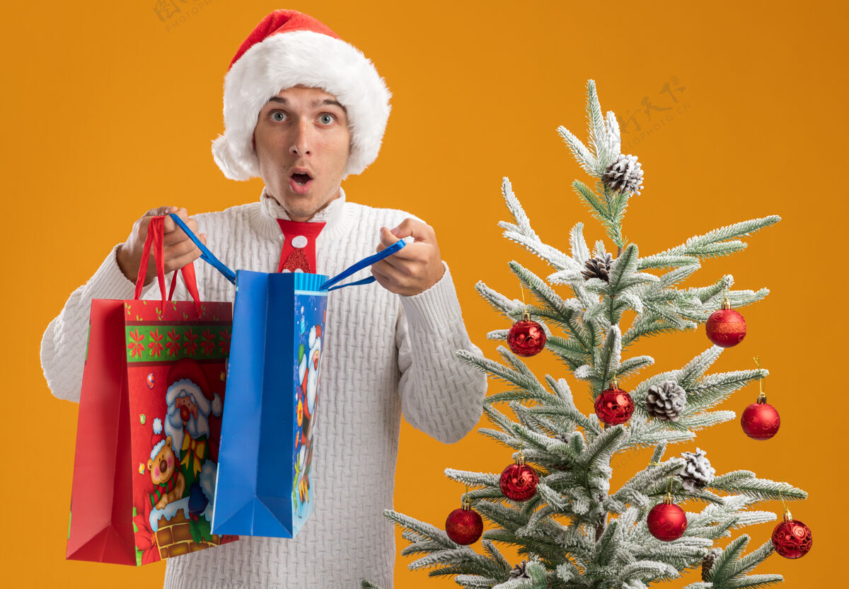 穿惊讶的年轻帅哥戴着圣诞帽 打着圣诞老人的领带 站在装饰好的圣诞树旁 手里拿着圣诞礼品袋 打开一个看起来孤零零的橙色墙上包领带小伙子