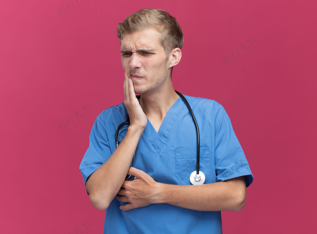 表情看着旁边年轻的男医生穿着医生制服用听诊器把手放在粉红色墙上孤立的疼痛的牙齿上牙齿制服壁板