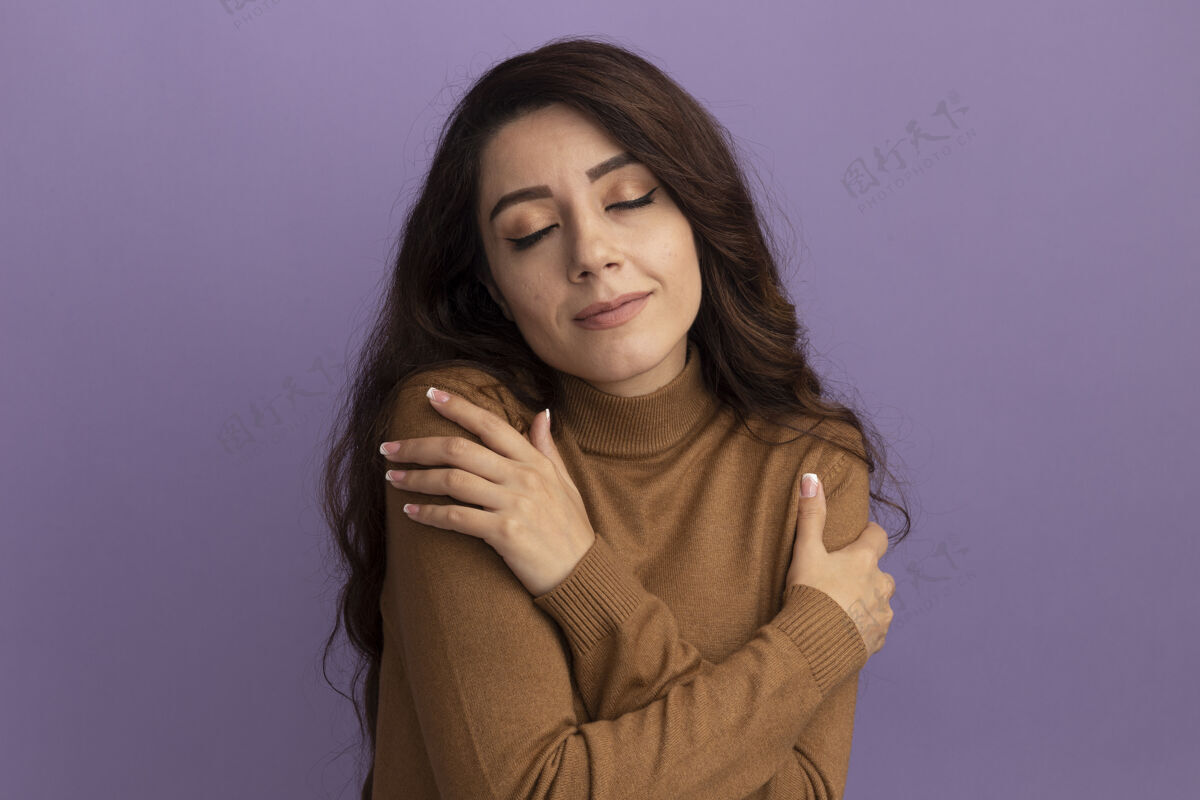 女孩喜欢闭上眼睛的年轻漂亮女孩穿着棕色高领毛衣把手放在肩膀上孤立在紫色的墙上衣服手封闭