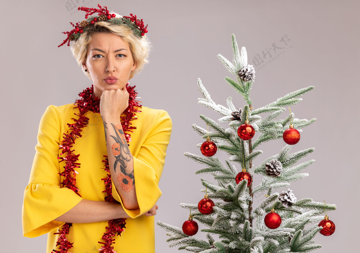 站严肃的金发女郎头戴圣诞花环 脖子上戴着金箔花环 站在装饰好的圣诞树旁 看着白色的墙上下巴下孤立的手保持花环花环