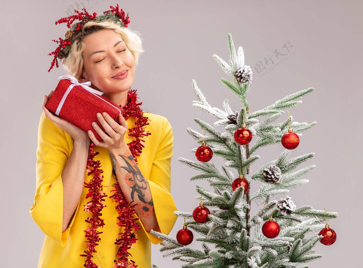 周围戴着圣诞花环 脖子上戴着金箔花环的年轻金发女郎站在装饰好的圣诞树旁 手里拿着礼品包 闭着眼睛孤立地站在白色的墙上请年轻近