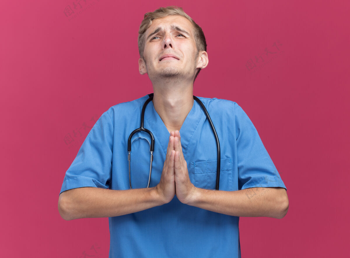 年轻人遗憾地看到年轻的男医生穿着医生制服 听诊器显示祈祷手势隔离在粉红色的墙上衣服站着男性