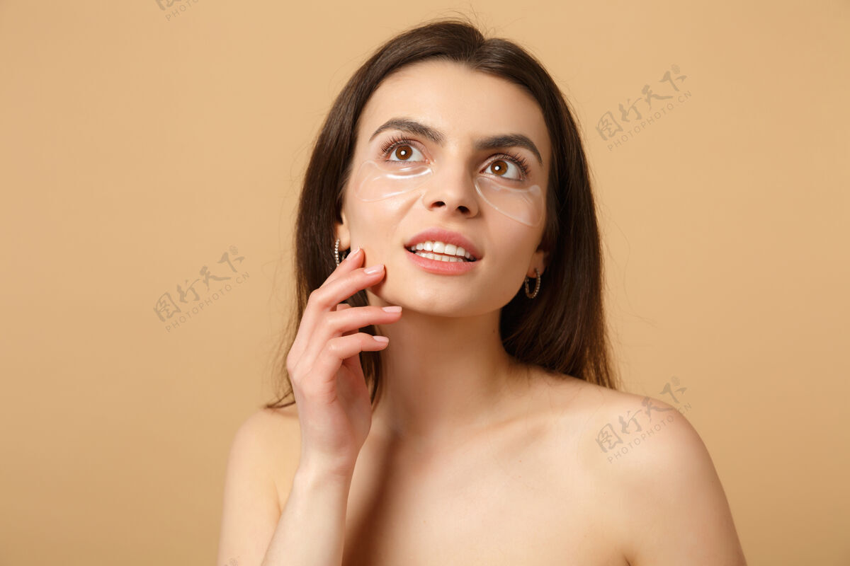 健康近距离拍摄完美皮肤的半裸女人 在米色粉彩墙上隔离眼睛下的裸妆贴片手治疗身体