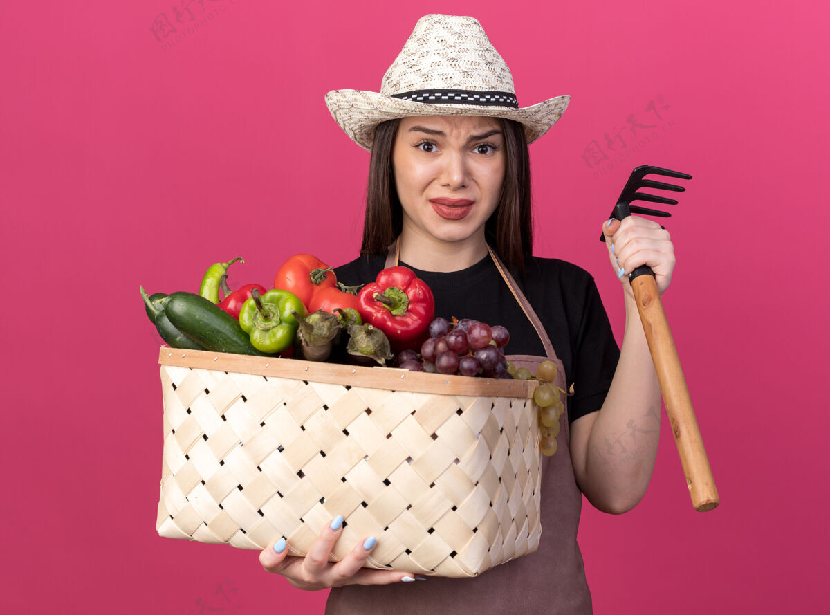 漂亮焦虑美丽的白人女园丁戴着园艺帽拿着菜篮和耙子举行耙帽子