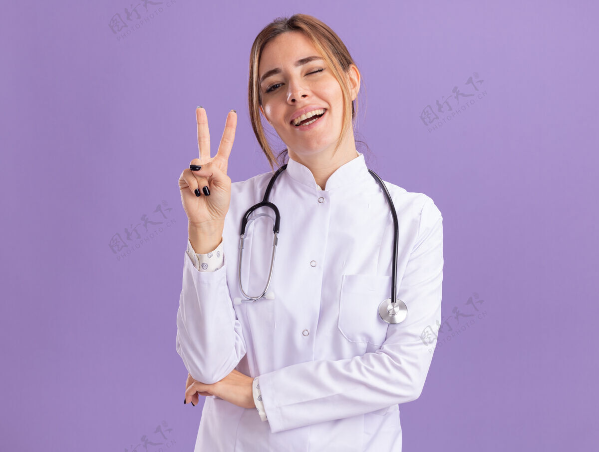 人年轻的女医生微笑着眨着眼睛 穿着医用长袍 听诊器显示出和平的姿态 隔离在紫色的墙上人穿着眨眼