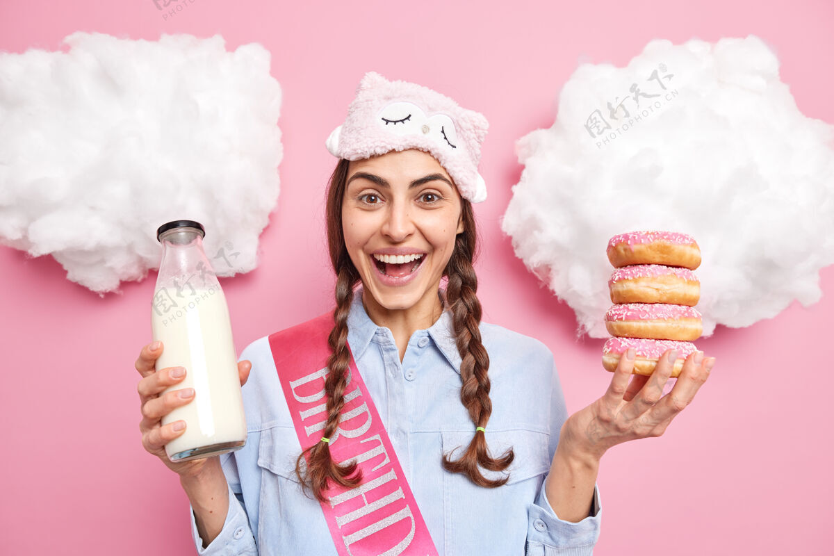 零食扎着两条辫子的女人微笑着 手里拿着美味的甜甜圈和牛奶 心情愉快 额头上戴着眼罩 隔离在粉色上室内穿食物