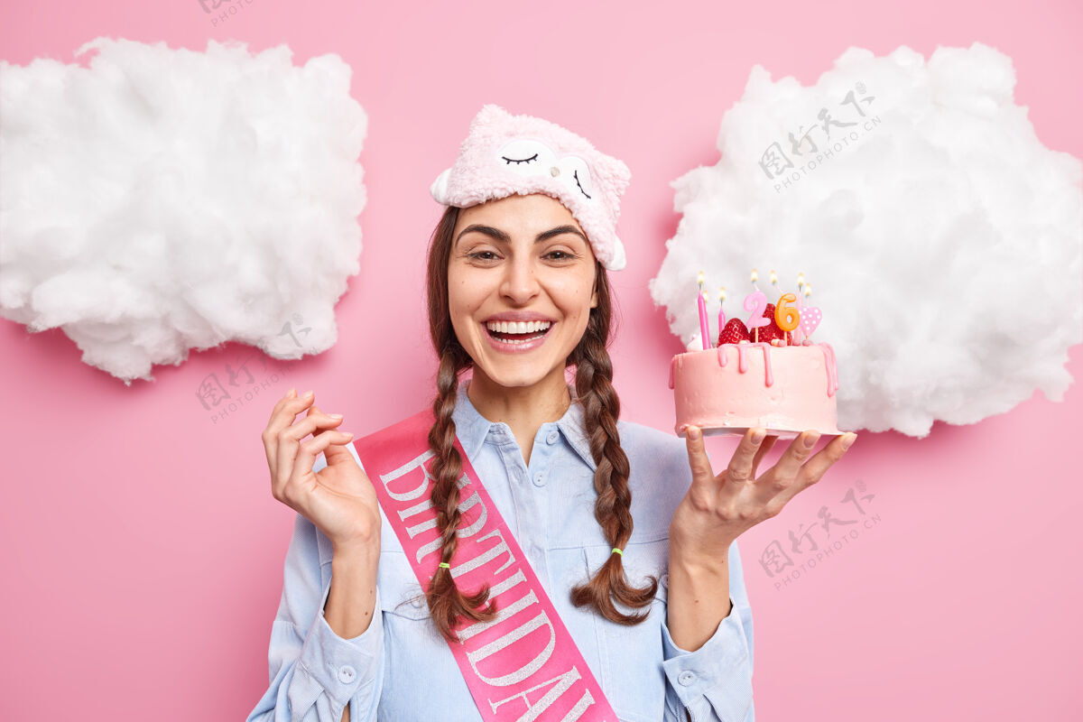 蛋糕女人开心地笑着拿着美味的草莓蛋糕和蜡烛庆祝生日享受家庭聚会戴着眼罩在额头上休闲衬衫隔离在粉红色的墙上生日积极庆典